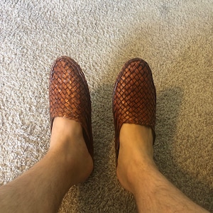 May include: A pair of brown leather woven slippers with a square toe. The slippers are worn on a person's feet.