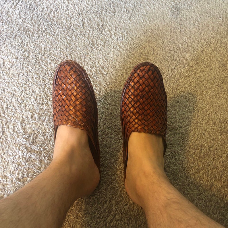 May include: A pair of brown leather woven slippers with a square toe. The slippers are worn on a person's feet.