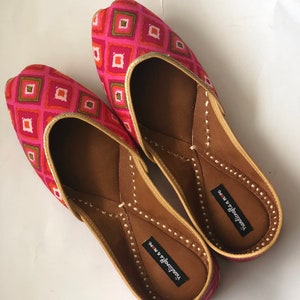 May include: A pair of pink and gold flat shoes with a geometric pattern. The shoes have a pointed toe and a small heel. The brand name "Handicrafts in Me" is printed on the inside of each shoe.