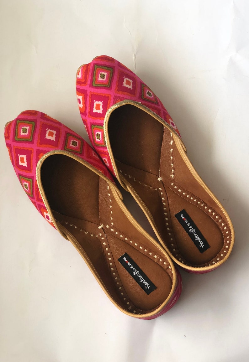 May include: A pair of pink and gold flat shoes with a geometric pattern. The shoes have a pointed toe and a small heel. The brand name "Handicrafts in Me" is printed on the inside of each shoe.