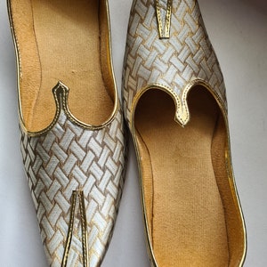 May include: A pair of gold and white woven fabric shoes with pointed toes. The shoes have a decorative gold trim and a pointed opening at the top.
