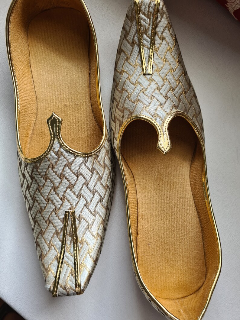 May include: A pair of gold and white woven fabric shoes with pointed toes. The shoes have a decorative gold trim and a pointed opening at the top.