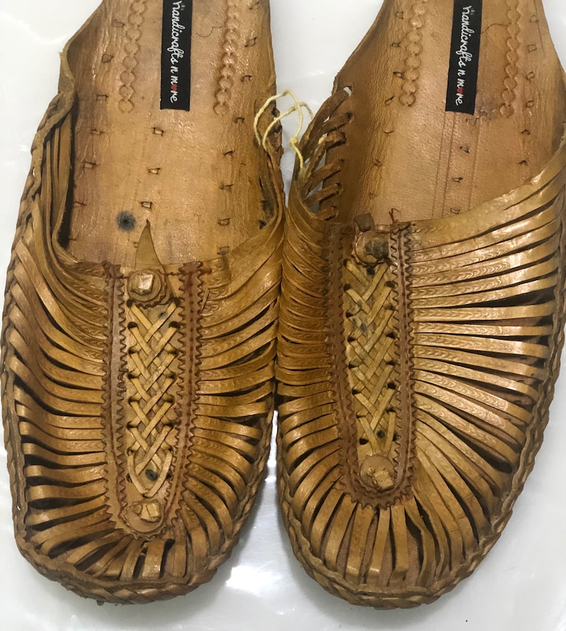 May include: A pair of tan leather sandals with intricate woven straps. The sandals have a simple design and are made of a soft, supple leather. The sandals are labeled "handicrafts n more".
