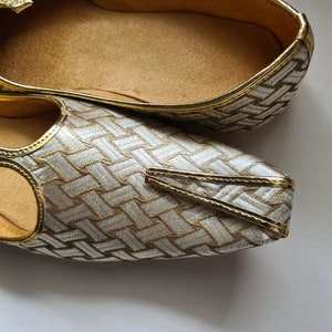 May include: A pair of gold and silver patterned shoes with a pointed toe and a decorative strap. The shoes have a gold trim and a soft, brown insole.