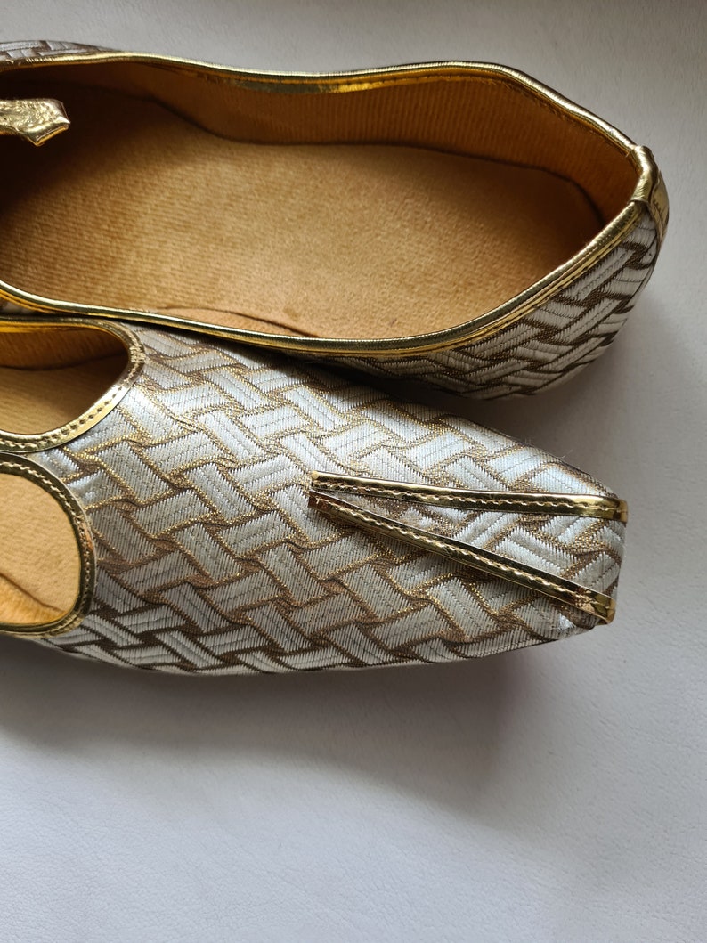 May include: A pair of gold and silver patterned shoes with a pointed toe and a decorative strap. The shoes have a gold trim and a soft, brown insole.