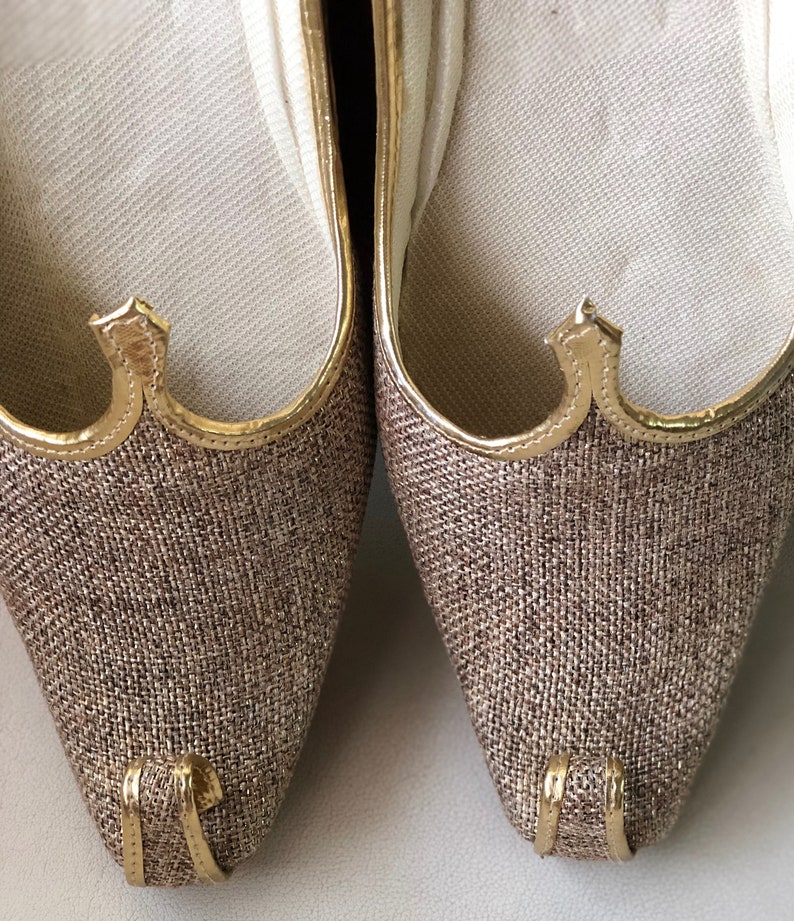 K&ouml;nnte beinhalten: Ein Paar braune und goldene spitze Schuhe mit einem strukturierten Obermaterial aus Stoff. Die Schuhe haben einen goldenen Besatz um die &Ouml;ffnung und einen goldenen Akzent an der Spitze.