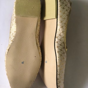 May include: A pair of gold woven flat shoes with a pointed toe. The shoes have a light brown sole with the word "al" written on the bottom.