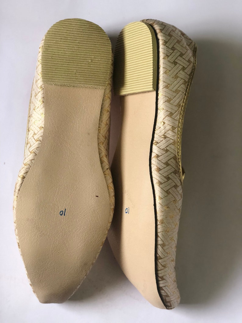 May include: A pair of gold woven flat shoes with a pointed toe. The shoes have a light brown sole with the word "al" written on the bottom.