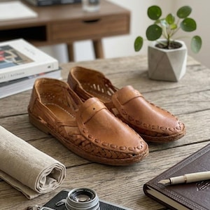 May include: A pair of tan leather loafers featuring a woven design across the vamp and sides, with a rounded toe, a low heel, and decorative stitching along the edges of the upper. The shoes have a slip-on construction with cut-out details.