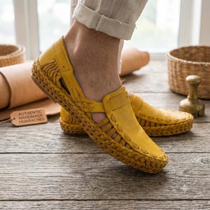 May include: A pair of yellow leather huarache shoes featuring a woven design, cut-out side panels, and a thick braided sole. A small rectangular tag nearby displays the text AUTHENTIC HANDMADE HUARACHES in black capital letters.