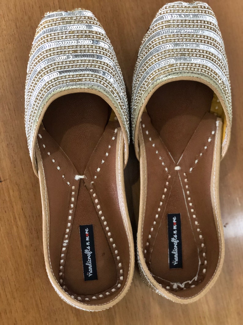 May include: A pair of gold and silver sequined flat shoes with a brown leather insole. The shoes have a pointed toe and a small strap across the top. The insole is labeled "handicrafts in me".