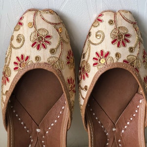 May include: A pair of cream-colored embroidered flat shoes with red and gold accents. The shoes have a pointed toe and a leather sole.