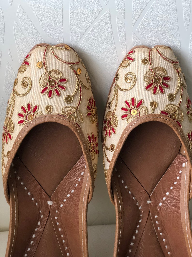 May include: A pair of cream-colored embroidered flat shoes with red and gold accents. The shoes have a pointed toe and a leather sole.