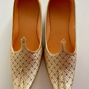 May include: A pair of gold-colored pointed-toe shoes with a woven pattern. The shoes have a pointed toe and a flat sole.