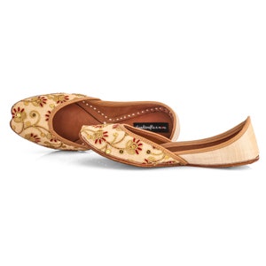 May include: A pair of beige embroidered flat shoes with red and gold floral designs. The shoes have a pointed toe and a low heel. The brand name "Handicrafts n more" is visible on the insole of one shoe.