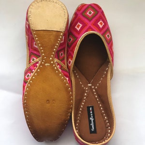 May include: A pair of pink and orange patterned flat shoes with leather soles. The shoes have a pointed toe and a small heel. The brand name "handicrafts n more" is printed on the inside of the shoe.