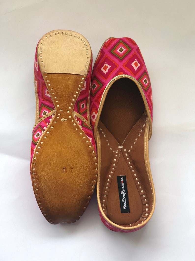 May include: A pair of pink and orange patterned flat shoes with leather soles. The shoes have a pointed toe and a small heel. The brand name "handicrafts n more" is printed on the inside of the shoe.