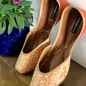 May include: A pair of peach-coloured embroidered flat shoes with gold thread. The shoes have a pointed toe and a small heel. The brand name "Youkoffee n me" is printed on the inside of the shoes.