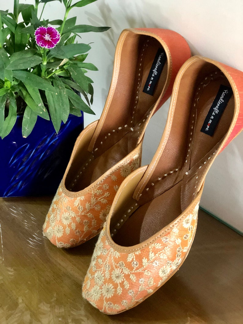 May include: A pair of peach-coloured embroidered flat shoes with gold thread. The shoes have a pointed toe and a small heel. The brand name "Youkoffee n me" is printed on the inside of the shoes.