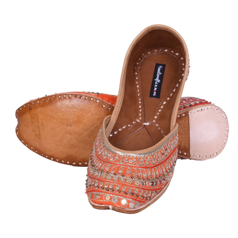 May include: A pair of orange and gold embroidered flat shoes with a leather sole. The shoes have a pointed toe and are decorated with sequins and beads. The brand name "Handicrafts & More" is visible on the inside of the shoe.