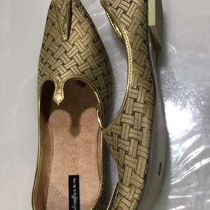 May include: A pair of gold woven flat shoes with a pointed toe and a small decorative piece on the top. The shoes have a label that reads "handicrafts n' me".