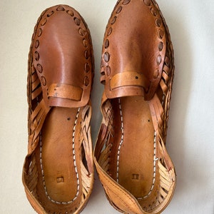 May include: A pair of tan leather huarache-style shoes featuring a woven side construction, a solid leather vamp with a decorative stitched border, and a flat sole with visible white stitching along the inner edge of the footbed.