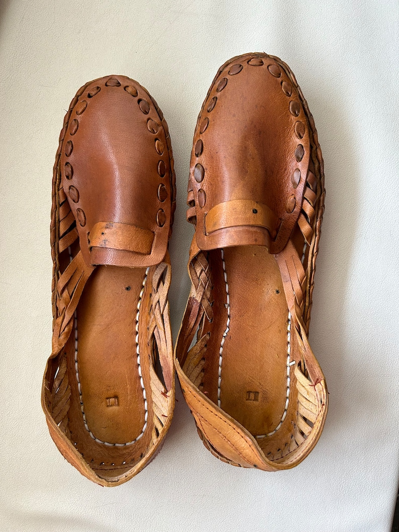 May include: A pair of tan leather huarache-style shoes featuring a woven side construction, a solid leather vamp with a decorative stitched border, and a flat sole with visible white stitching along the inner edge of the footbed.