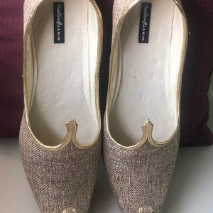 K&ouml;nnte beinhalten: Ein Paar goldene, glitzernde, spitz zulaufende flache Schuhe mit goldener Verzierung. Die Schuhe haben eine wei&szlig;e Innensohle mit dem in Schwarz gedruckten Text "handicrafts in home".