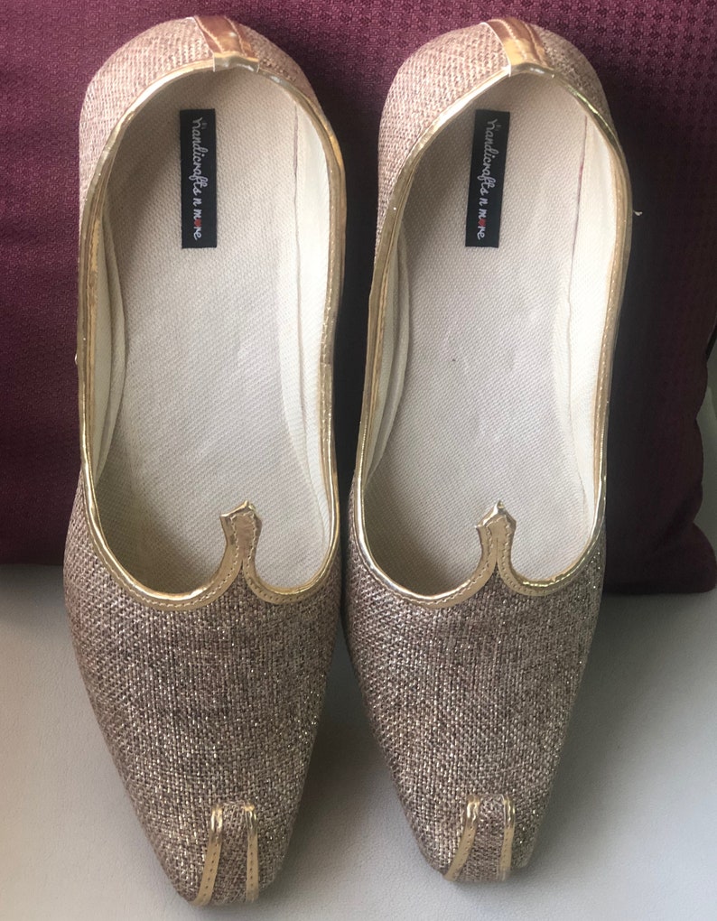 K&ouml;nnte beinhalten: Ein Paar goldene, glitzernde, spitz zulaufende flache Schuhe mit goldener Verzierung. Die Schuhe haben eine wei&szlig;e Innensohle mit dem in Schwarz gedruckten Text "handicrafts in home".