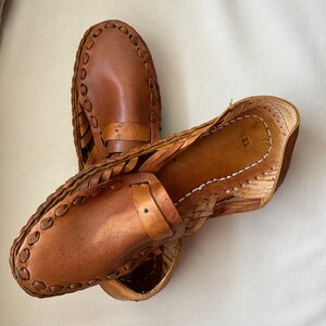 May include: A pair of tan leather loafers featuring a woven side construction and a decorative strap across the vamp. The shoes have a rounded toe, visible stitching along the edges, and a smooth leather finish with a natural light brown color.