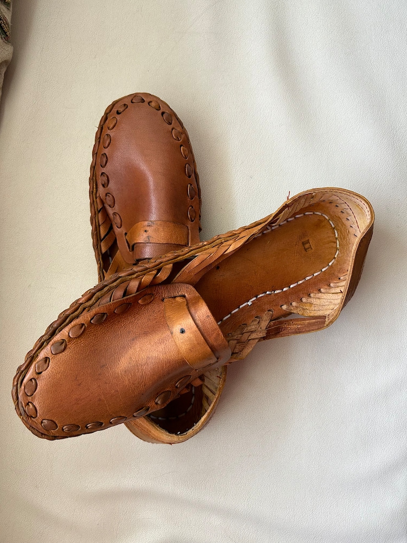 May include: A pair of tan leather loafers featuring a woven side construction and a decorative strap across the vamp. The shoes have a rounded toe, visible stitching along the edges, and a smooth leather finish with a natural light brown color.