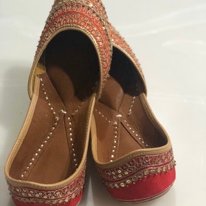 May include: A pair of red and gold embroidered flat shoes with pointed toes. The shoes have a traditional Indian design and are decorated with sequins and beads.