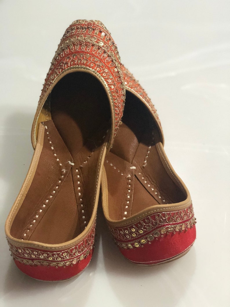 May include: A pair of red and gold embroidered flat shoes with pointed toes. The shoes have a traditional Indian design and are decorated with sequins and beads.
