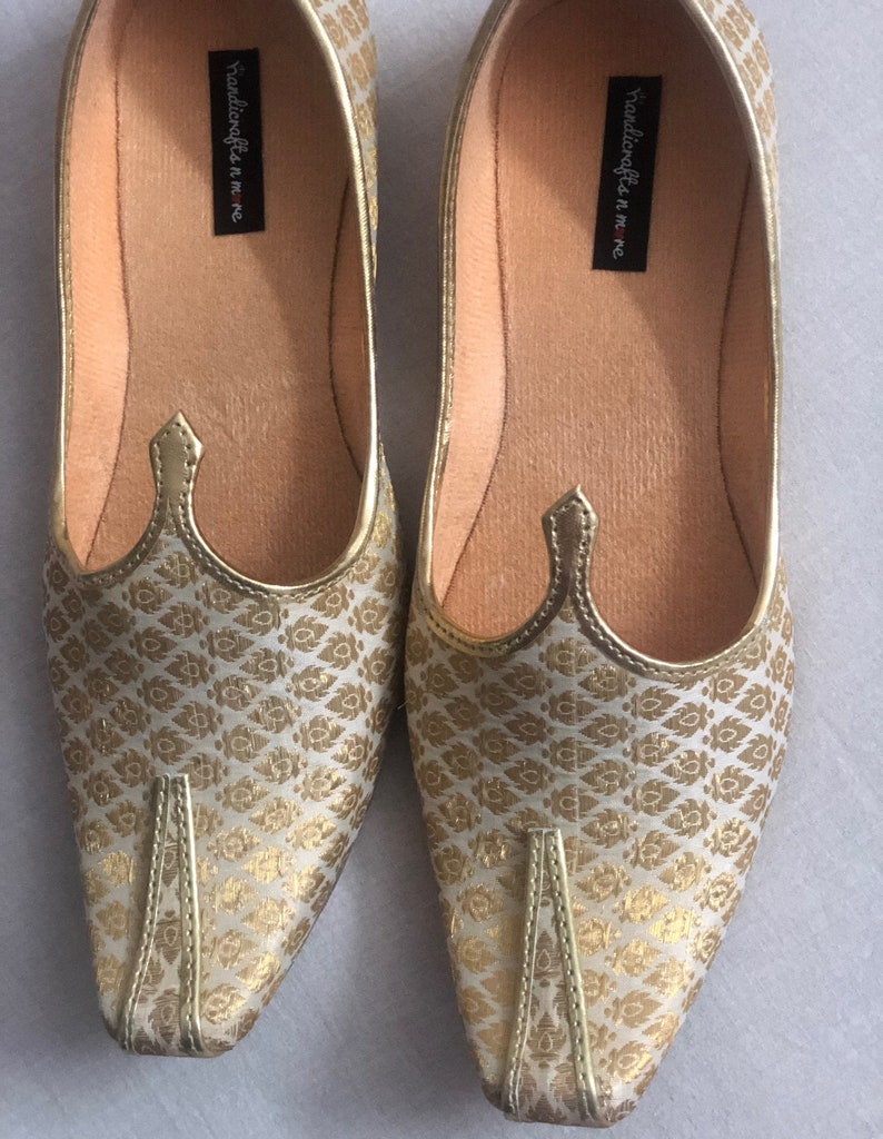 May include: A pair of gold and white patterned flat shoes with pointed toes. The shoes have a decorative detail on the top and the words "handicrafts in me" are printed on the inside of the shoes.