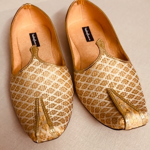 May include: A pair of gold and white patterned flat shoes with a pointed toe. The shoes have a decorative strap across the top and the brand name "sandalya.in" is printed on the inside of the shoes.