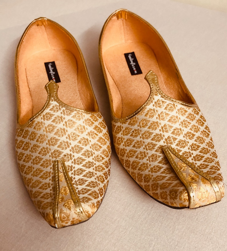 May include: A pair of gold and white patterned flat shoes with a pointed toe. The shoes have a decorative strap across the top and the brand name "sandalya.in" is printed on the inside of the shoes.