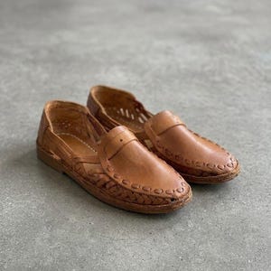 May include: A pair of tan leather huarache shoes featuring a woven design across the toe and sides, a closed heel, and a flat sole. The leather has a smooth finish with visible stitching details along the edges and a strap across the vamp.