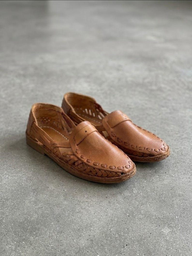 May include: A pair of tan leather huarache shoes featuring a woven design across the toe and sides, a closed heel, and a flat sole. The leather has a smooth finish with visible stitching details along the edges and a strap across the vamp.