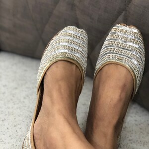 May include: A pair of gold and silver embellished flat shoes. The shoes have a pointed toe and a low heel. The embellishments are arranged in a horizontal pattern.