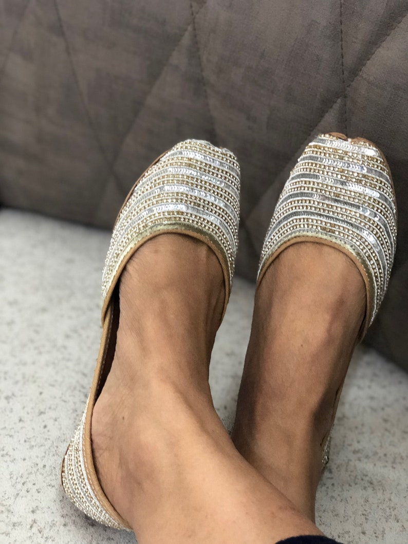May include: A pair of gold and silver embellished flat shoes. The shoes have a pointed toe and a low heel. The embellishments are arranged in a horizontal pattern.