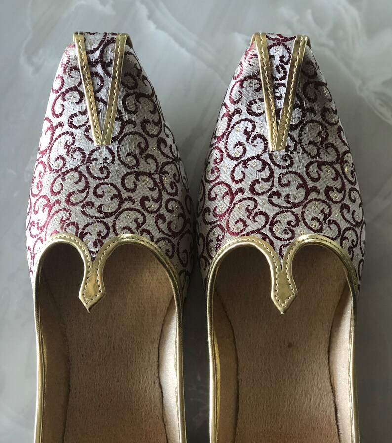 May include: A pair of white and burgundy embroidered shoes with gold trim. The shoes have a pointed toe and a closed back.