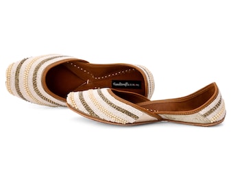 May include: White and gold beaded flat shoes with a brown leather sole. The shoes have a pointed toe and a low heel. The brand name "handicrafts n more" is printed on the side of the shoe.