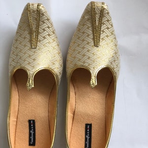 May include: A pair of gold and white woven fabric shoes with a pointed toe and a decorative strap. The shoes have a cream-colored lining and the text "handicrafts n more" is printed on the inside of each shoe.