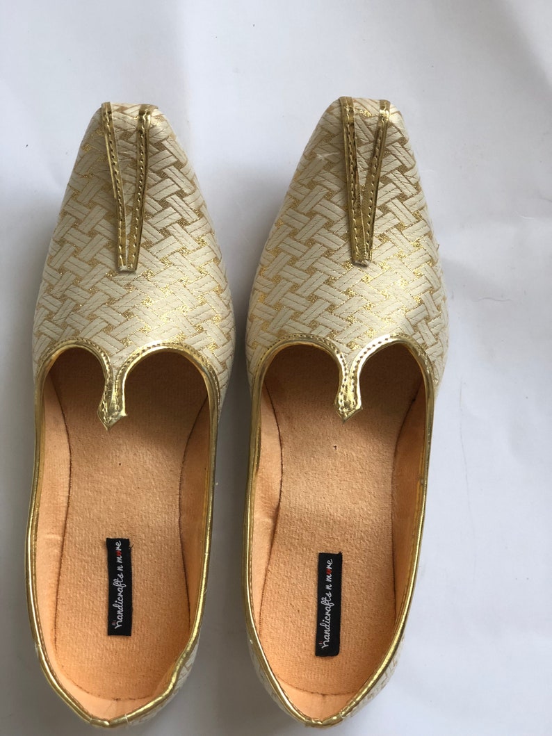 May include: A pair of gold and white woven fabric shoes with a pointed toe and a decorative strap. The shoes have a cream-colored lining and the text "handicrafts n more" is printed on the inside of each shoe.