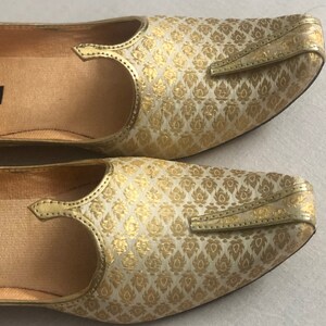 May include: A pair of gold and white patterned flat shoes with a pointed toe and a decorative strap.