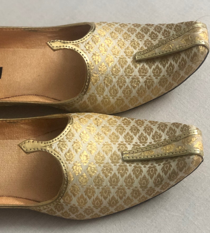 May include: A pair of gold and white patterned flat shoes with a pointed toe and a decorative strap.