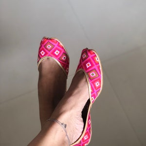 May include: A pair of pink and gold embroidered flat shoes with a pointed toe. The shoes have a geometric pattern and are worn on a person's feet.