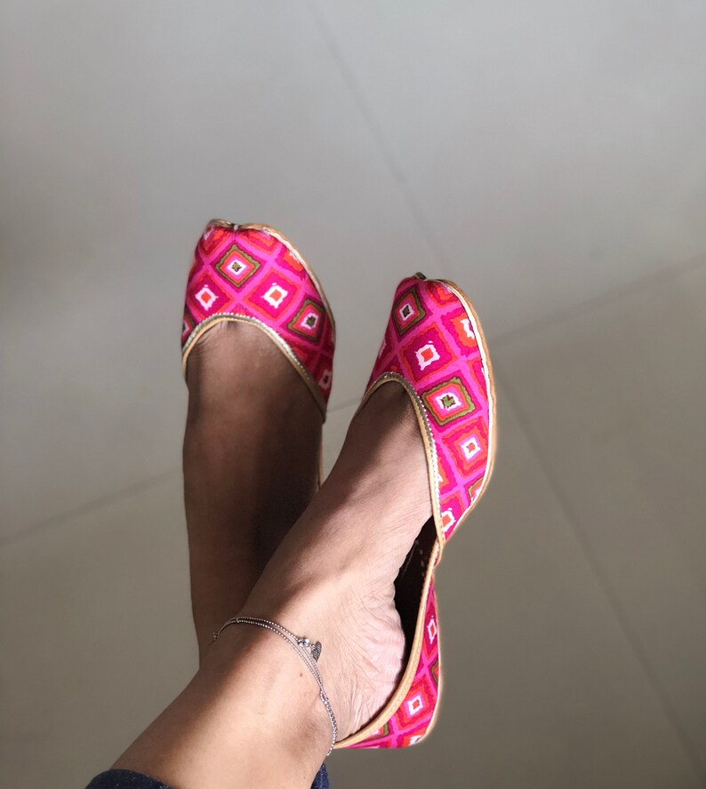 May include: A pair of pink and gold embroidered flat shoes with a pointed toe. The shoes have a geometric pattern and are worn on a person's feet.
