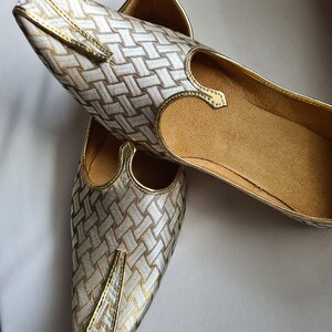 May include: A pair of gold and white woven fabric shoes with pointed toes. The shoes have a decorative strap across the top.