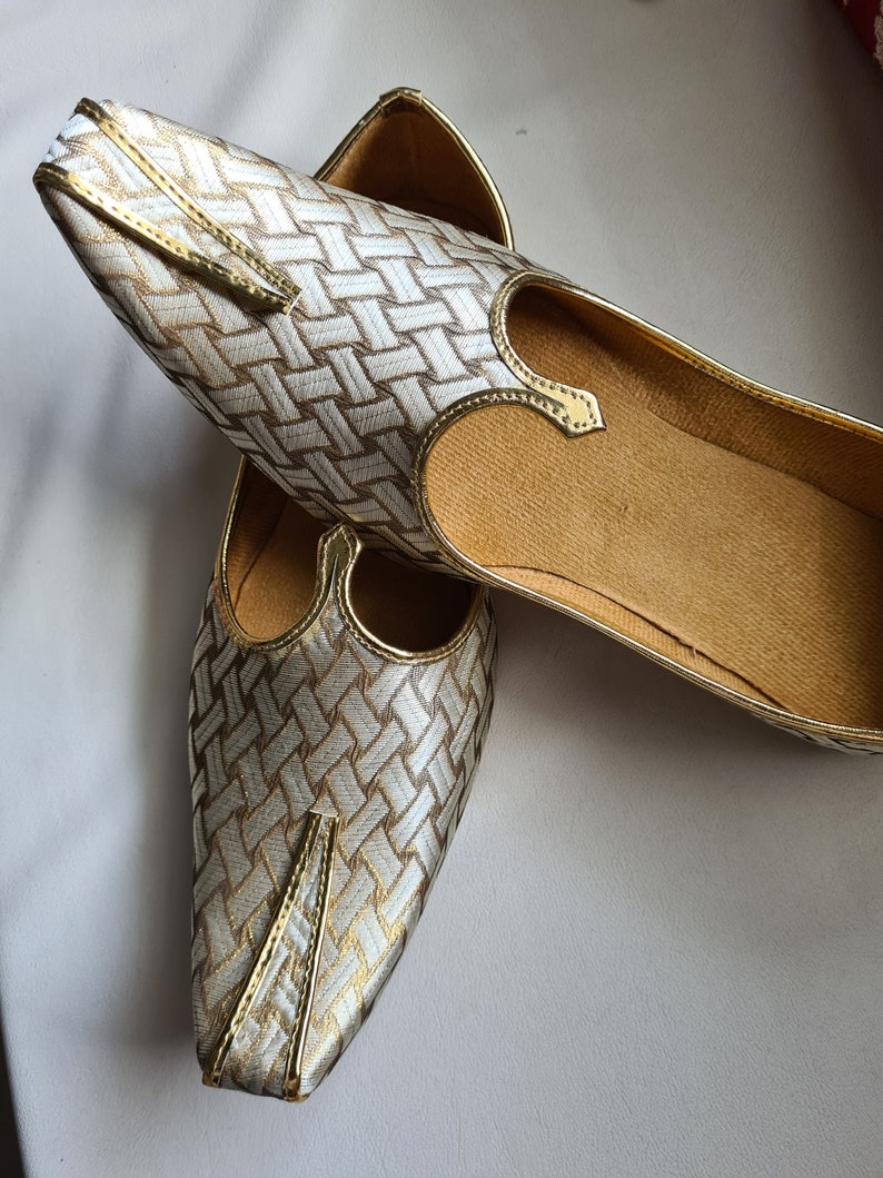 May include: A pair of gold and white woven fabric shoes with pointed toes. The shoes have a decorative strap across the top.